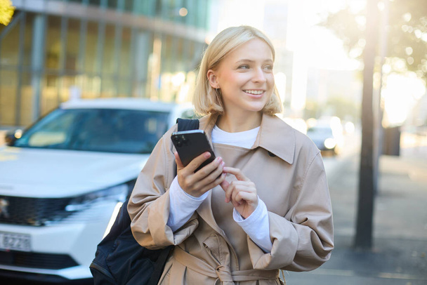 Portrait of young woman using map application on smartphone, student with backpack, walks along street on sunny day, holds mobile phone in hand. - Photo, Image