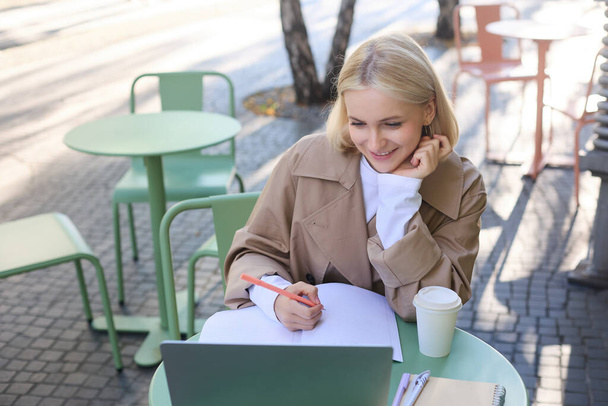 Imagen de una joven estudiante sonriente, trabajando en el proyecto, sentada afuera en el día soleado en la cafetería, mirando a la computadora portátil y tomando notas, estudiando, haciendo tareas al aire libre. - Foto, imagen