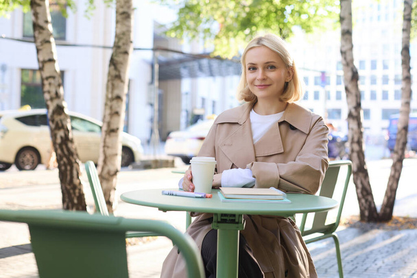 Image of young woman, makes sketches outdoors on street in notebook, drinks coffee and smiles at camera. Lifestyle and people concept - Photo, Image