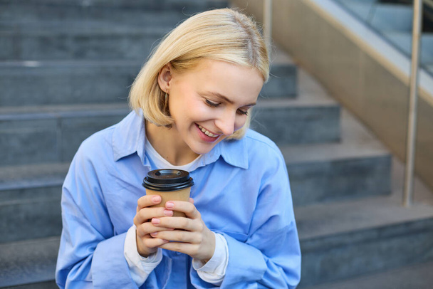 Nahaufnahme Porträt einer schönen, lächelnden blonden Frau, Studentin, die auf einer Treppe außerhalb des Campus sitzt, Kaffee zum Mitnehmen trinkt, ihre Hände wärmt, während sie eine Tasse hält. - Foto, Bild
