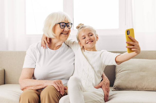Selfie white child hugging grandmother two smiling phone photography family people sofa call bonding togetherness t-shirt granddaughter space video copy education indoors - Фото, изображение