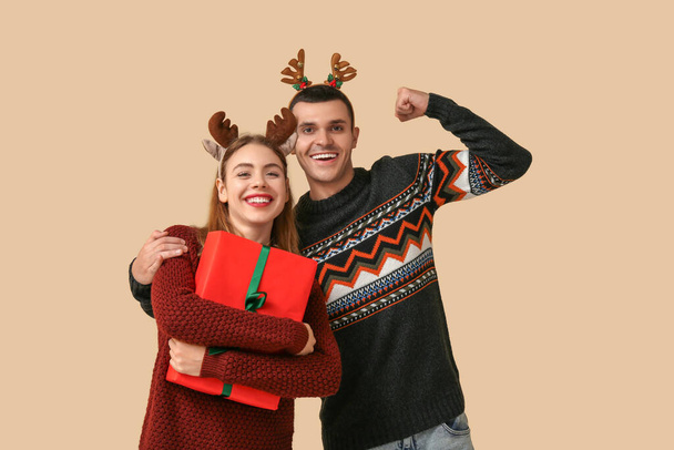 Beau jeune couple en cornes de rennes tenant boîte cadeau de Noël et montrant les muscles sur fond beige - Photo, image
