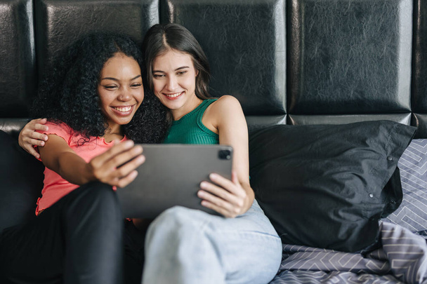 LGBT kadınları yatakta oturup tablet ekranı izlerken mutlu bir şekilde gülümsüyor ve evde tatil aktivitelerinde birlikte yaşıyorlar. - Fotoğraf, Görsel