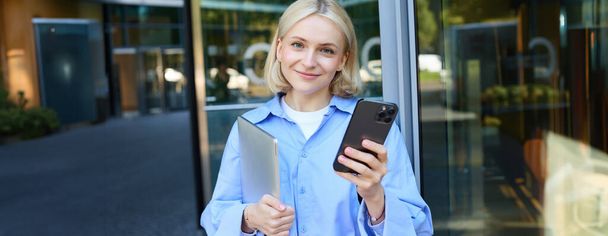 Lebensstilaufnahme einer jungen lächelnden Frau, Studentin oder Angestellten in der Nähe eines Bürogebäudes, Handy und Laptop in den Händen haltend, glücklich blickend, Benachrichtigung auf dem Smartphone prüfend. - Foto, Bild