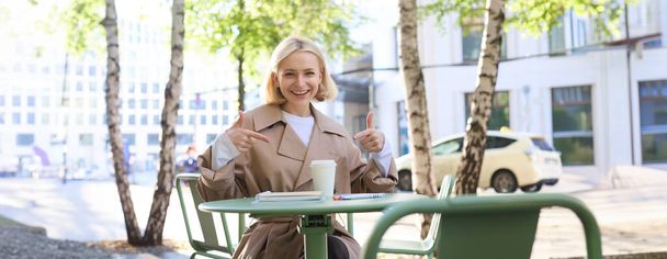 Fröhlich lächelnde junge Frau sitzt im Café, zeigt mit dem Finger auf Kaffeetasse zum Mitnehmen, empfiehlt guten Außenraum, nickt zustimmend und sieht zufrieden aus. - Foto, Bild