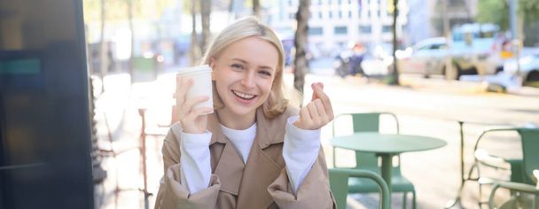 Gente y concepto de ocio. Chica rubia feliz, sentado con una taza de café en la cafetería de la calle, mostrando el signo del corazón del dedo, disfruta pasar tiempo al aire libre. - Foto, Imagen
