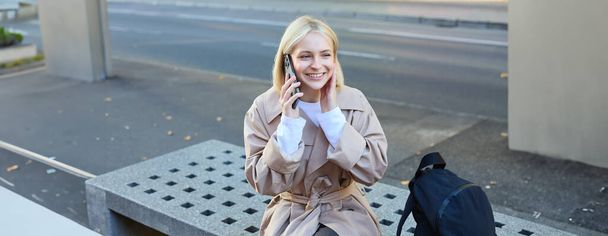 Imagen de una joven rubia sentada en el banco de la calle con una mochila, hablando por teléfono móvil, responde a una llamada y sonríe mientras conversa con alguien. - Foto, imagen