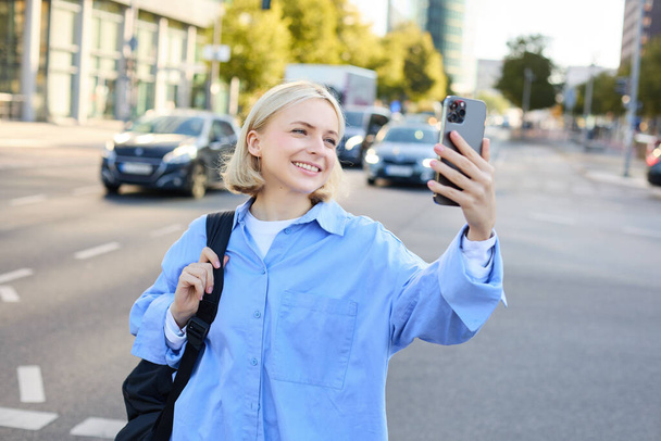 Изображение улыбающейся молодой женщины, современной девушки со смартфоном, делающей селфи, снимающей фото оживленных улиц города, записывающей видео на открытом воздухе. - Фото, изображение