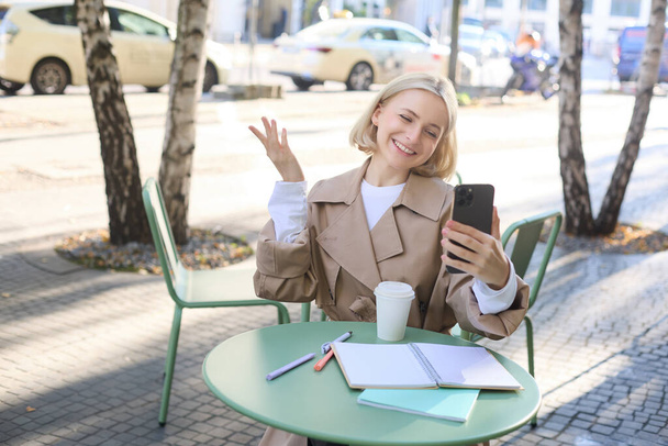 Jeune mannequin blonde, étudiante dans un café, discuter de quelque chose au téléphone, bavarder vidéo, regarder la caméra du smartphone et sourire, parler à un ami via une application mobile. - Photo, image