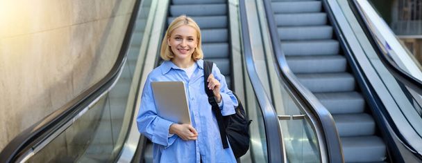 Retrato de una joven modelo femenina con portátil y mochila en el hombro, posando cerca de la escalera mecánica, mirando a la cámara con cara sonriente confiada. - Foto, Imagen