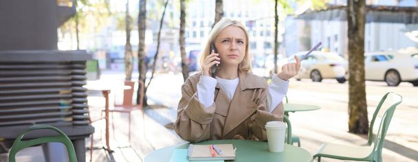Портрет женщины, студентки, делающие домашнее задание на улице в кафе, держащие ручку, хмурящиеся во время звонка кому-то, пытающиеся понять телефонный разговор. - Фото, изображение