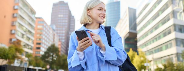 Gros plan portrait de la jeune femme blonde élégante, debout dans la rue, vérifier son téléphone mobile, en utilisant l'application de smartphone pour se déplacer en ville, en regardant la carte en ligne pour les directions. - Photo, image