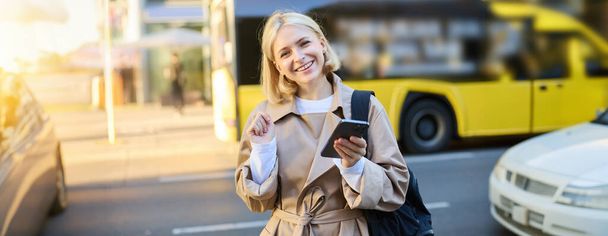 Street style shot of smiling young blond woman, holding mobile phone, standing on street and looking happy, holding backpack and wearing trench coat. - Photo, Image