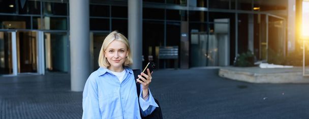 Retrato vertical de jovem gerente de escritório, mulher de colarinho azul camisa e mochila, segurando laptop e smartphone, esperando perto de edifício de negócios na rua, sorrindo para a câmera. - Foto, Imagem