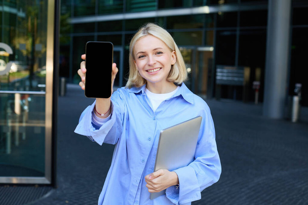 Arckép elegáns, modern fiatal irodai munkás, nő mutatja neki smartphone képernyő, mobiltelefon app kijelzőn, pózol közelében campus vagy irodaház, gazdaság laptop. - Fotó, kép