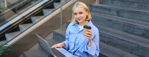 Wizerunek stylowej młodej nowoczesnej kobiety, studentki odrabiającej lekcje, studiującej na zewnątrz na schodach kampusu, siedzącej z laptopem i kawą, pijącej jej cappuccino i łączącej się z ogólnodostępnym WiFi. - Zdjęcie, obraz