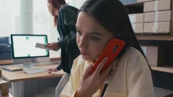 Keskikokoinen kuva naispuolisesta ylläpitäjästä puhumassa matkapuhelimella asiakkaiden kanssa, kun taas toinen työntekijä pakkaamassa laatikoita varastointipaikassa markkinoilla - Materiaali, video