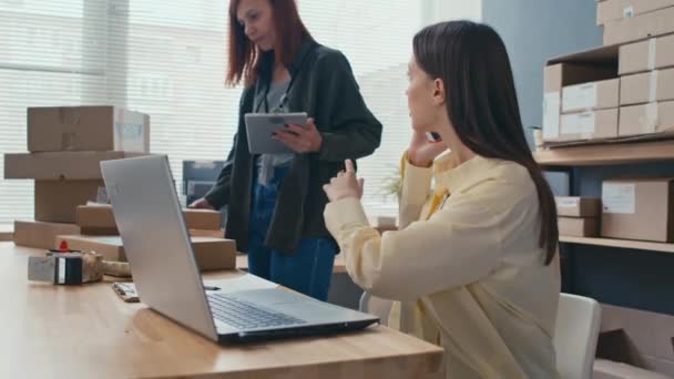 Μεσαία λήψη του θηλυκού διευθυντή προετοιμασία κουτιά με παραγγελίες για τους πελάτες, ενώ ο συνάδελφός της μιλάει στο τηλέφωνο με τους πελάτες και τη χρήση φορητού υπολογιστή - Πλάνα, βίντεο