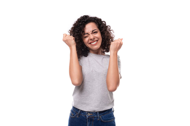 kind slender young caucasian woman with careless black curly hair is dressed in a gray t-shirt on a white background. - Photo, Image