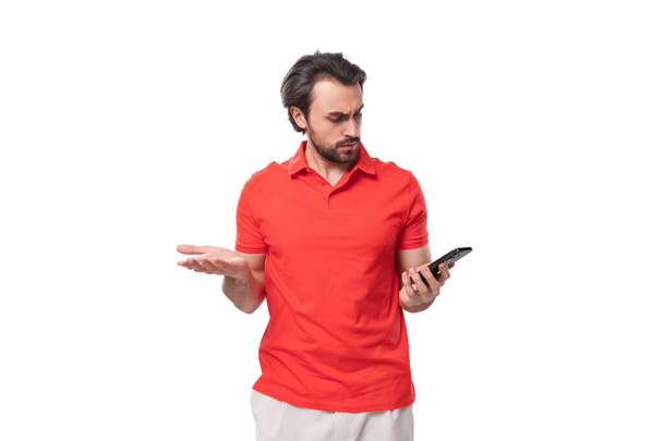 mladý pohledný muž s černými vlasy a vousy oblečený v červeném tričku vrhá ruce pomocí smartphonu. - Fotografie, Obrázek