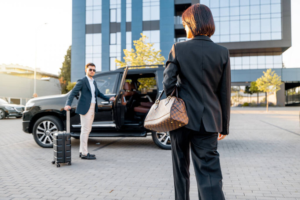 Elegancka bizneswoman wchodzi do luksusowego samochodu, mężczyzna z walizką otwiera drzwi samochodu, wpuszcza kobietę. Pojęcie podróży służbowej i podróży - Zdjęcie, obraz