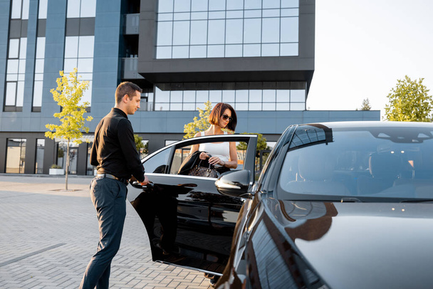 Şoför zarif bir iş kadınının arabaya binip ofis binasının ya da otelin yanındaki özel bir taksinin kapısını açmasına yardım ediyor. Kişisel sürücü kavramı veya iş seyahatleri - Fotoğraf, Görsel