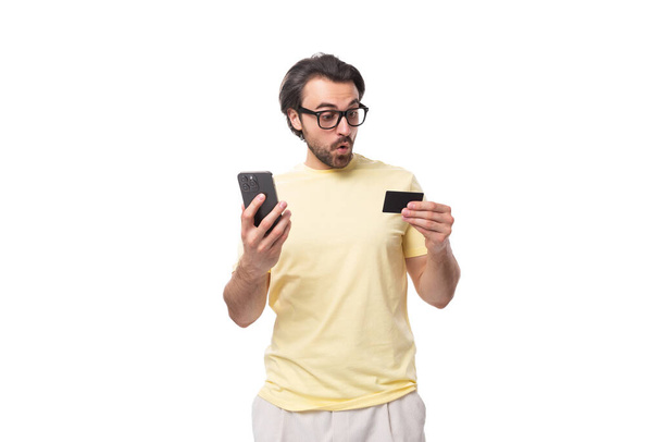 tipo caucásico casual con una barba en gafas vestidas con una camiseta ligera sostiene un teléfono y una tarjeta de crédito. - Foto, Imagen