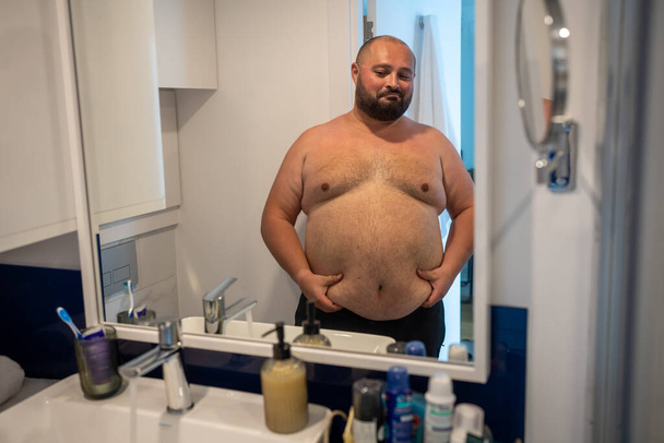 Χοντρός άντρας κοιτάζει τον καθρέφτη χαμογελώντας αγγίζοντας μεγάλη κοιλιά στο μπάνιο στο σπίτι. Ο γενειοφόρος δεν ικανοποιήθηκε με το σχήμα του σώματός του. παχυσαρκία, υπέρβαρο αρσενικό, επιπλέον βάρος, υγιή προβλήματα, έλεγχος βάρους. - Φωτογραφία, εικόνα