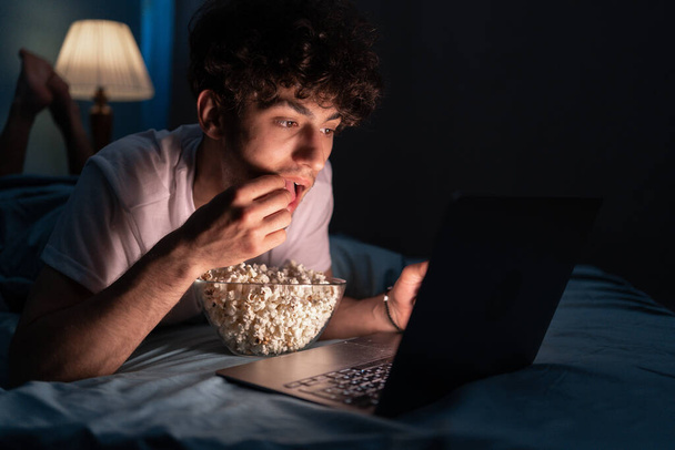 Szczęśliwy muzułmanin jedzący popcorn leżąc w łóżku oglądając film na laptopie w nocy w domu. Przestrzeń kopiowania - Zdjęcie, obraz
