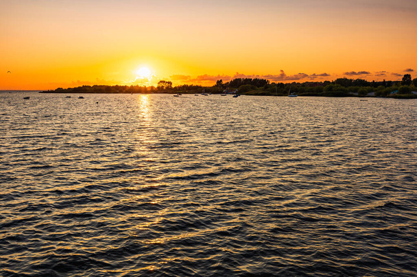 Schöner wolkenverhangener Sonnenuntergang über dem Meer mit ein paar Booten und Kitesurfern und ein paar Bäumen und Büschen neben kleinem Strand - Foto, Bild