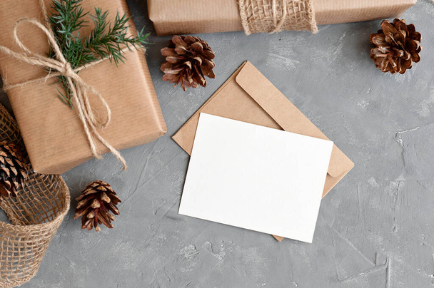 エレガントな持続可能なクリスマスの装飾とプレゼント,ギフトタグ,ポストカードモックアップ. 空の紙カード,作られたギフト用の箱,松のツイッグ,灰色のテーブルの背景のコーン. ホリデー冬の招待テンプレート. - 写真・画像