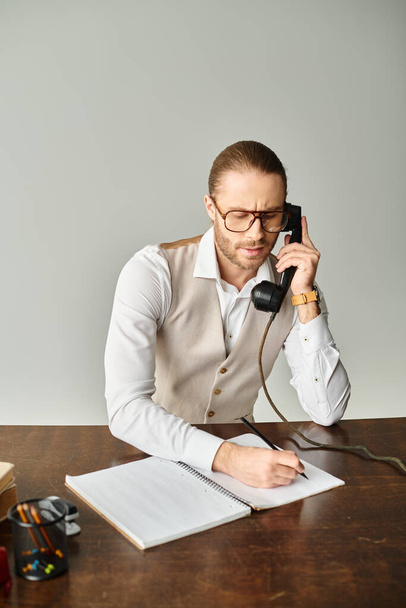 schöner bärtiger Mann mit Brille, der per Retro-Telefon spricht und sich am Tisch im Büro Notizen macht - Foto, Bild