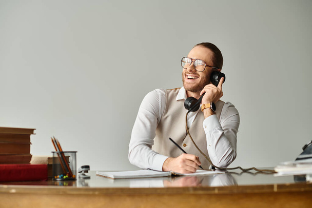 wesoły młody człowiek z brodą i okularami rozmawiający przez retro telefon i robiący notatki, uśmiechający się szczęśliwie - Zdjęcie, obraz