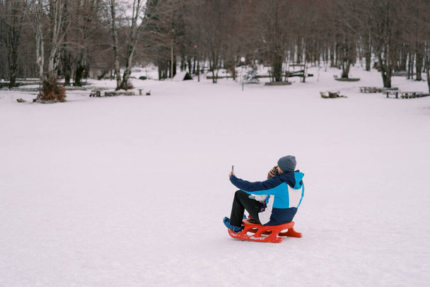 Pai tirando selfie smartphone de si mesmo e criança sentada no trenó no gramado nevado. Vista traseira. Foto de alta qualidade - Foto, Imagem