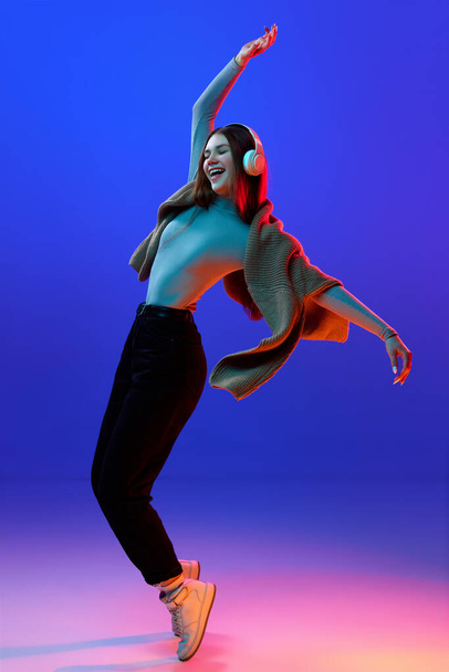 Overjoyed, happy girl, student dancing while listening music in headphones against blue studio background in neon light, filter. Concept of beauty, human emotions, hobby, fashion and style. Ad - Fotoğraf, Görsel