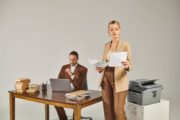atrakcyjna młoda kobieta w eleganckim garniturze patrząc na papierkową robotę obok jej chłopaka pracującego na laptopie - Zdjęcie, obraz