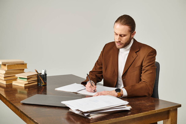 όμορφος νεαρός άνδρας με γενειάδα σε καφέ σακάκι κάθεται στο γραφείο και κρατώντας σημειώσεις προσεκτικά - Φωτογραφία, εικόνα