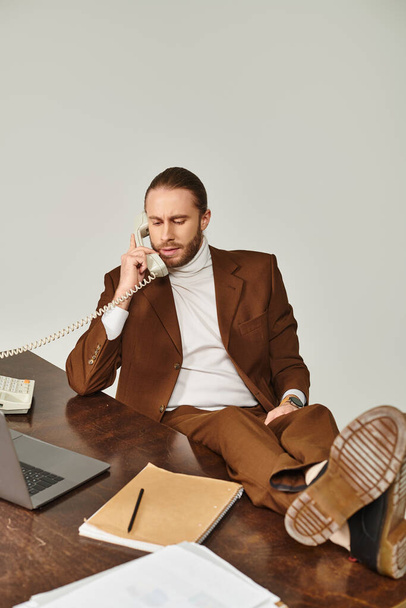 привлекательный трудолюбивый мужчина с бородой в коричневом костюме разговаривает по ретро-телефону с ногами на столе - Фото, изображение