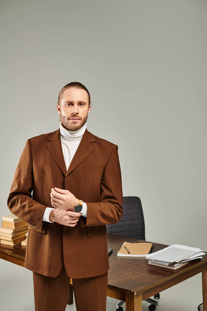 knappe bebaarde man in modieuze bruine jas kijkend naar camera op grijze achtergrond door tafel - Foto, afbeelding