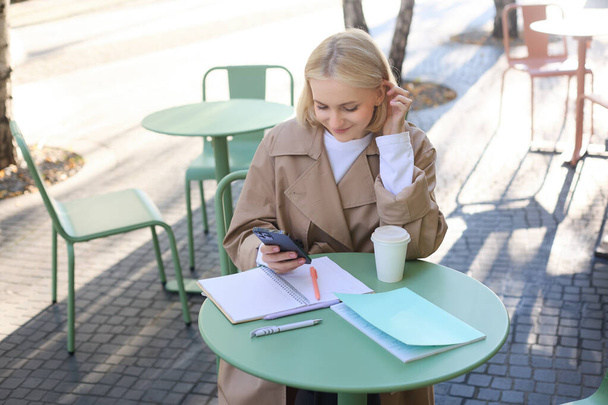 Obraz młodej pięknej dziewczyny, student studiuje, odrabianie pracy domowej w kawiarni, przy użyciu telefonu komórkowego, trzymając smartfona, uśmiechnięty. - Zdjęcie, obraz