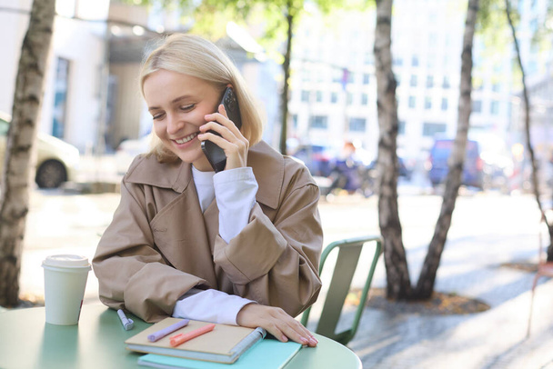 Πορτρέτο της νεαρής όμορφης γυναίκας στο καφέ, κάθεται σε εξωτερικούς χώρους τη φωτεινή ηλιόλουστη μέρα, μιλώντας στο smartphone, κουβεντιάζοντας από το τηλέφωνο και χαμογελώντας. - Φωτογραφία, εικόνα