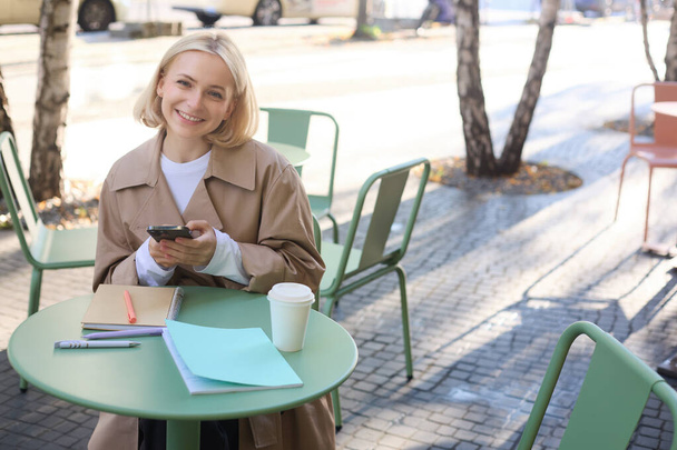 Retrato de una linda mujer sonriente con el pelo corto rubio, sentado con documentos y teléfono móvil, mirando feliz a la cámara. - Foto, imagen