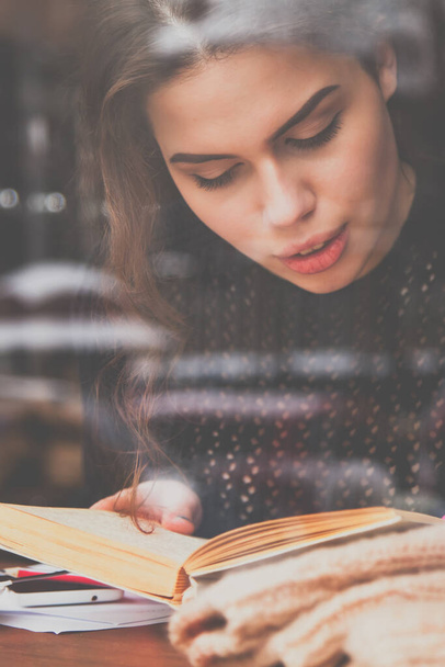 Портрет красивой молодой женщины читает книгу в кафе в холодный зимний день. Вид за стеклом. Отдых, учеба, знания, концепция успеха. Горизонтальное изображение. - Фото, изображение