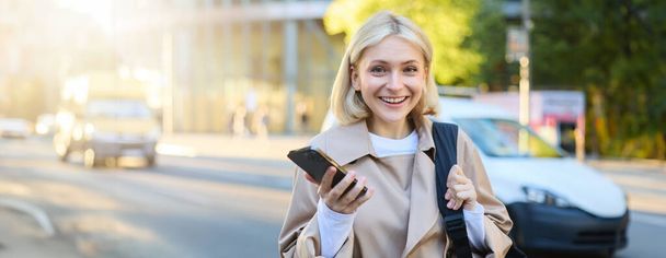 Close up portret młodej nowoczesnej kobiety, stojącej na ulicy, student uniwersytetu z telefonem komórkowym, szczęśliwy i uśmiechnięty do kamery, za pomocą aplikacji smartphone map. - Zdjęcie, obraz