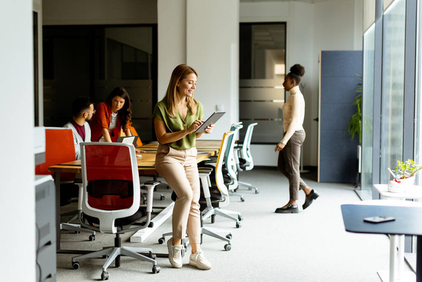 Νεαρή γυναίκα στέκεται με ψηφιακή πλακέτα μπροστά στην ομάδα της στο σύγχρονο γραφείο - Φωτογραφία, εικόνα