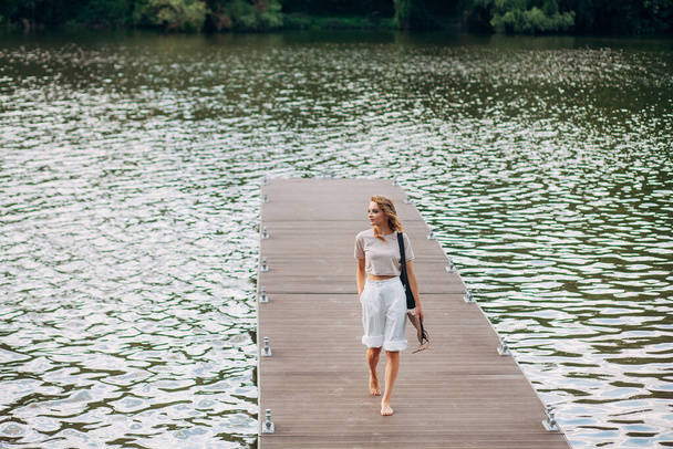 Een mooi meisje met modieuze make-up en kapsel, in witte broek en een beige top, met een zwarte leren handtas, loopt langs een bootpier op een pittoreske rivier. - Foto, afbeelding