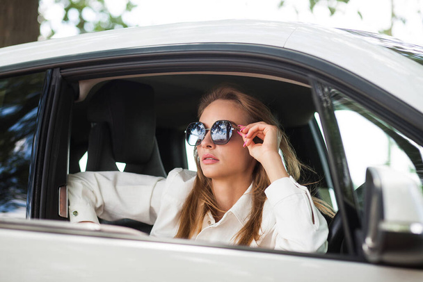 Módní krásná štíhlá blondýnka ve slunečních brýlích, světlých šortkách a vestě sedí v černobílém autě. Obrázek pro módní bloggery. - Fotografie, Obrázek