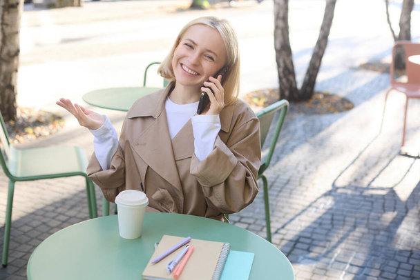 Outgoing young smiling woman, sitting in outdoor cafe, drinking coffee, answer phone call, talking on mobile, chatting with friendly attitude on smartphone. - Photo, Image
