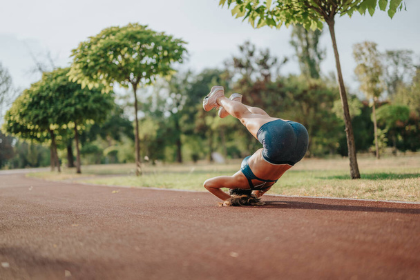 Houkutteleva urheilullinen tyttö suorittaa headstand ulkona vihreässä puistossa, jossa on joustavuutta, voimaa ja tasapainoa. Hänen istuva ruumiinsa ja inspiroiva ympäristö ilmentävät aktiivisen urheilun kauneutta. - Valokuva, kuva