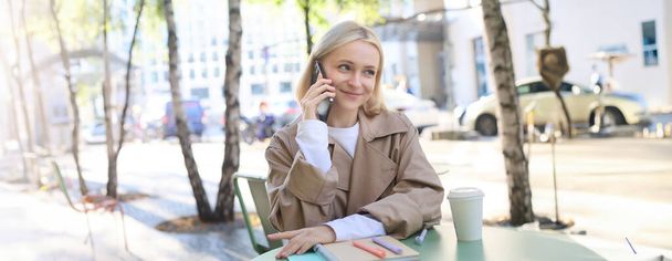 Zbliżenie portret uśmiechniętej blondynki, siedzącej przy stole przed kawiarnią, rozmawiającej przez telefon komórkowy, mającej szczęśliwą, ożywioną rozmowę. - Zdjęcie, obraz